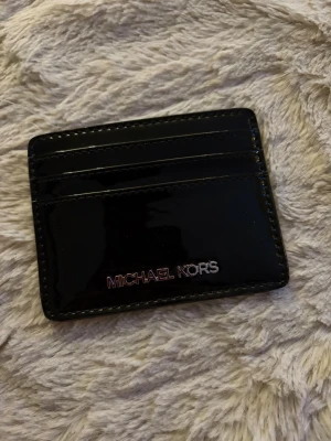 Svart kortfodral Michael Kors - Snyggt svart kortfodral från Michael Kors i glansigt material. Fodralet har flera kortfack på ena sidan och ett genomskinligt fack på baksidan, perfekt för ID eller busskort. Stilren och smidig design som passar i alla fickor. Ny oanvänd köptes i present box med väska , men använder inte korthållare så därav säljer jag den 