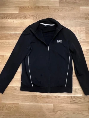  Hugo Boss Hoodie - Stilren svart hoodie med dragkedja och vita detaljer från Boss Hugo Boss. Utrustad med hög krage och diskret logotyp på bröstet, perfekt kombination av komfort och design.