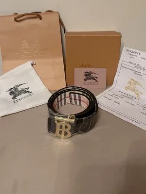 Vändbart bälte (beige/svart) med guldlogga - Exklusivt vändbart bälte med två sidor: beige mönstrad sida samt svart för en mer diskret look. Guldlogga som ger en elegant och lyxig touch. Ett tidlöst accessoarval som lyfter vilken outfit som helst.