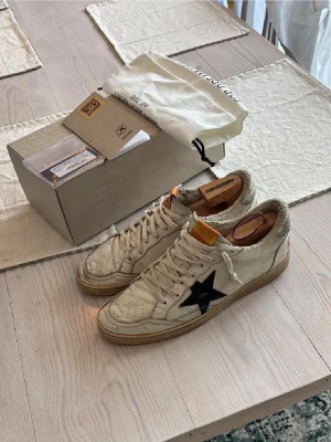 Golden Goose ballstars skor - Säljer ett par ikoniska Golden Goose sneakers i vitt skinn med svart stjärna på sidan och röda detaljer baktill. Skorna har snörning, perforerade detaljer på tån och en beige sula. Tungan har en orange etikett med GGDB-logga. Råa kanter runt öppningen ger en cool, avslappnad vibe.