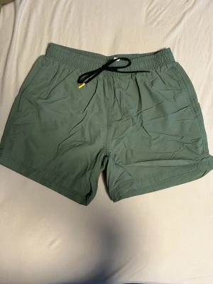Gröna shorts med snörning - Snygga gröna shorts med elastisk midja och svart snörning med gul detalj. De har en enkel design med två sidofickor och är tillverkade i ett lätt syntetmaterial, perfekta för varma dagar eller strandhäng. Kommer stryka innan jag skickar!
