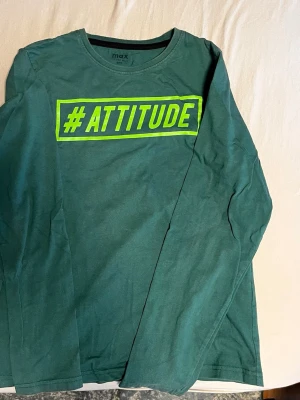 Grön långärmad tröja med tryck - Cool grön långärmad tröja från max med stort neongult tryck på bröstet där det står #ATTITUDE. Rund halsringning och avslappnad passform, perfekt för dig som vill sticka ut lite extra.