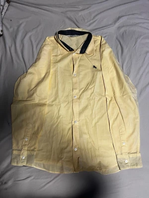 Gul skjorta från H&M med kontrastkrage - Gul skjorta från H&M i bomull med mörkblå kontrastkrage och broderad detalj på bröstet. Klassisk knappstängning framtill och långa ärmar med manschett. Perfekt för dig som vill sticka ut lite extra med en färgglad look.