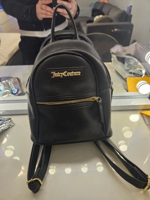 Svart ryggsäck från Juicy Couture - Säljer en svart miniryggsäck från Juicy Couture med guldfärgad logga framtill och dragkedja. Väskan är i syntetmaterial och har justerbara axelband. Insidan är fodrad med mönstrat tyg med Juicy Couture-tryck. Perfekt för dig som vill ha en snygg och praktisk accessoar. Är köpt i usa. Pris kan diskuteras! 