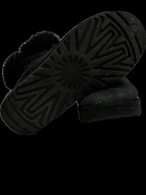 Svarta UGG boots med fårskinn - Mysiga svarta UGG boots med fluffigt fårskinnsfoder och dekorativ knapp på sidan. Skorna har en platt sula och är tillverkade i mjuk mocka. Perfekta för kalla dagar när du vill hålla fötterna varma och sköna.