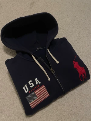 Ralph Lauren Hoodie - Fräsch och exklusiv Ralph Lauren zip Hoodie med en stor röd ponny och USA logga. Storleken är L och skicket är helt perfekt, använd max 3 gånger. Ny pris: 2000kr+, Mitt pris 799kr. Hör av dig vid frågor!😊✨