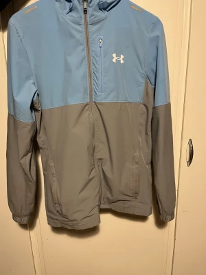 Blå och grå vindjacka från Under Armour - Snygg tvåfärgad vindjacka från Under Armour i ljusblått och grått. Jackan har huva, dragkedja framtill, bröstficka med zip och diskret logga på bröstet. Perfekt för dig som vill ha en sportig look och gillar funktionella detaljer. Använd endast två gånger och ser nästintill ny ut. Jackan är bekväm, lätt och passar perfekt för både vardag och träning. Säljes eftersom den inte kommer till användning.