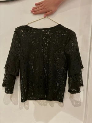 Svart spetsblus med volangärm - Säljer en svart blus i genombruten spets med trekvartsärm och volangdetaljer. Blusen har en elegant och feminin look med transparent material och blommönstrad spets. Perfekt att styla med jeans eller kjol för en trendig outfit.
