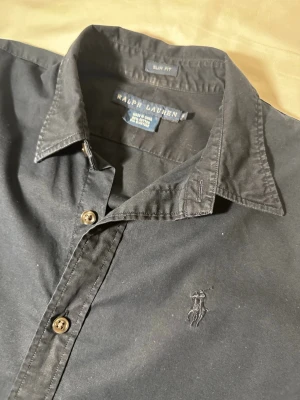 Svart slim fit skjorta från Ralph Lauren - Svart skjorta från Ralph Lauren i slim fit-modell med klassisk krage och knappar framtill. Materialet är mjuk bomull med lite stretch (95% bomull, 5% elastan). Diskret broderad logga på bröstet och långa ärmar med knappslå. Perfekt för en clean och stilren look.