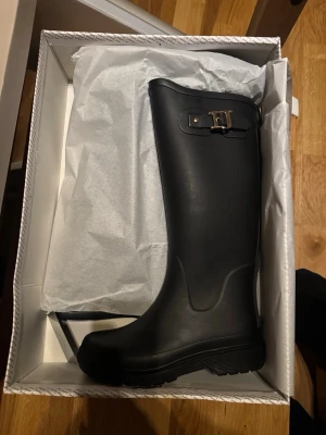 Svarta gummistövlar Ilse Jacobsen - Svarta höga gummistövlar från Ilse Jacobsen med snyggt spänne upptill. Klassisk design med rund tå och grov sula, perfekta för regniga dagar. Tillverkade i slitstarkt syntetmaterial som håller fötterna torra.