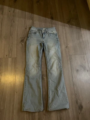 Ljusblå bootcut jeans med broderi - Säljer ett par ljusblå jeans med bootcut passform och snyggt rosa broderi på bakfickorna. Två knappar och dragkedja framtill. Jeansen är mjuka och sköna att ha på sig. Använda fåtal gånger.