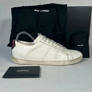Vita sneakers från Saint Laurent - Säljer ett par klassiska vita sneakers från Saint Laurent med diskret logga på plösen och röd detalj på hälen. Skorna har rund tå, snörning och är tillverkade i slätt skinn. Perfekta för dig som gillar stilrena och tidlösa sneakers. Dom kommer med original boxen, påsen och extra sko snören 