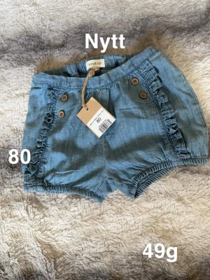 Blå volangshorts från Newbie - Söta blå shorts från Newbie med volangdetaljer och träknappar framtill. Shortsen har resår i midjan och benen för skön passform. Perfekta för varma dagar och lätta att matcha med andra plagg.🌸 Ny med lapp. Storlek 80