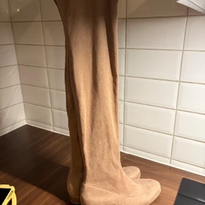 Beiga mockastövlar med låg klack - Säljer ett par höga stövlar i beige mocka med rund tå och låg klack. Stövlarna har en stilren design utan synliga detaljer och passar perfekt till  jeansMaterialet är mjukt och ger en lyxig känsla. Dom kommer över knäna 