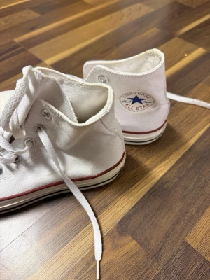 Vita Converse All Star high tops - Klassiska vita Converse All Star high tops med röd rand runt sulan och vita snören. Skorna är i textil med rund tå och platt sula. Converse-logga på sidan och metallhål för snörningen. Perfekta för dig som gillar tidlös streetstil.