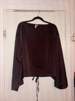 Brun omlottopp från Nelly - Säljer en brun omlottopp från Nelly i storlek 34. Toppen har långa ärmar, rund halsringning och knytband i midjan som ger en snygg siluett. Materialet är glansigt och känns lyxigt, perfekt att styla med jeans eller kjol.