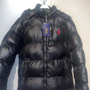 Svart pufferjacka från Polo Ralph Lauren - Svart pufferjacka från Polo Ralph Lauren med huva och dragkedja framtill. Jackan har ett rött broderat logomärke på bröstet och två sidofickor med dragkedja. Materialet är glansigt och känns riktigt chill för kalla dagar. Står storlek M men passar S 
