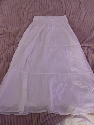 Vit långkjol med resårmidja - Säljer en vit långkjol med bred resår i midjan. Kjolen har en luftig och flowy känsla med flera sömmar som ger ett snyggt fall. Perfekt för varma dagar och enkel att matcha med olika toppar.