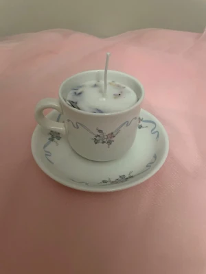 Doftljus av vaniljdoft i en vit tekopp med blommor och blåa streck nr 5 - Det här ljuset är gjort av återanvändbara material. Tekoppen är köpt secondhand och stearinet är från kyrkor som tänkte slänga det. Det här ljuset har en svag doft utav vanilj, så känsliga även kan använda det. Det här ljuset har även torkade blommor på som dekoration. Det finns fler av denna design på hemsidan om man vill köpa sets. 