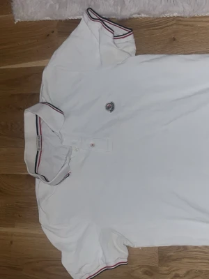 Vit Moncler pikétröja  - Vit pikétröja från Moncler med klassisk krage och knappar framtill. Kragen och ärmsluten har snygga ränder i rött och marinblått. Tröjan har Moncler-logga broderad på bröstet och är gjord i mjuk bomull. Perfekt för en clean och sportig look.