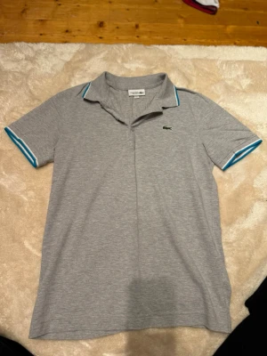 Grå pikétröja från Lacoste Sport - Snygg grå pikétröja från Lacoste Sport med klassisk krage och korta ärmar. Tröjan har blå och vita detaljer på krage och ärmslut samt den ikoniska gröna krokodilloggan på bröstet. Perfekt för en sportig och stilren look.