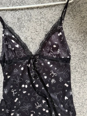 Svart långklänning Miss Selfridge - Supersöt svart långklänning från Miss Selfridge med vita hjärtan, rosetter och körsbärsmönster. Klänningen har smala axelband, spetsdetaljer vid urringningen och nederkanten samt snörning framtill. Materialet är glansigt och känns lätt mot huden.