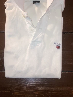 Vit polo piké från GANT i bomull - Säljer en klassisk vit polo piké från GANT i storlek S. Toppen har krage, knappar framtill och broderad GANT-logga med sköld på bröstet. Tillverkad i 100% bomull för en skön och luftig känsla. Perfekt för dig som gillar stilrena och tidlösa plagg.