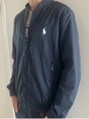 Mörkblå vindjacka från Ralph Lauren - Säljer denna vind/regn jacka från Ralph Lauren i storlek XL men passar M. Jackan har aldrig använts och har kvar originaletiketterna. Väldigt stilren, speciellt till kyligare sommarkvällar samt även fram mot hösten.