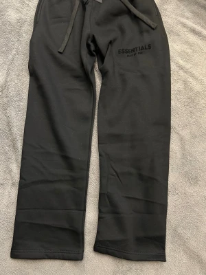 Svarta Essentials Fear of God mjukisbyxor - Svarta mjukisbyxor från Essentials Fear of God med broderad logga på benet och patch vid midjan. Byxorna har snörning i midjan och är tillverkade i mjukt bomullsmaterial. Perfekt för chill dagar och streetwear-stil.