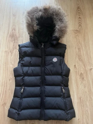 Svart dunväst med päls Moncler - Säljer en svart dunväst från Moncler med fluffig huva kantad av päls. Västen har två dragkedjefickor, quiltad design och klassisk Moncler-logga på bröstet. Perfekt för kyliga dagar när du vill vara både varm och snygg.
