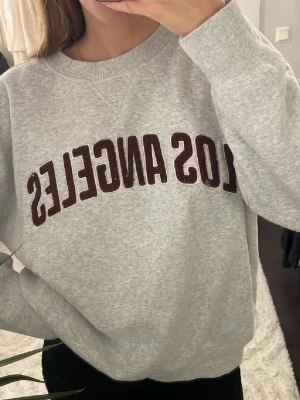 Grå sweatshirt från H&M  - Säljer en grå sweatshirt med mörkrött 'Los Angeles'-tryck på bröstet. Tröjan har rund hals, långa ärmar och ribbade muddar. Perfekt för en chill och avslappnad stil. Materialet känns mjukt och skönt mot huden.