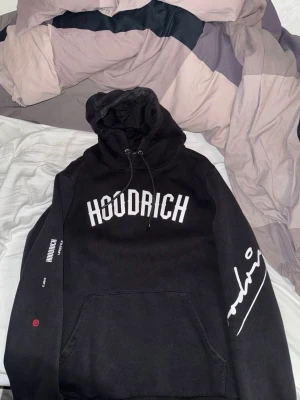 HOODRICH hoodie - Svart hoodie från HOODRICH med stor vit logga på bröstet och tryck på ärm och rygg. Klassisk huva med snörning och känguruficka framtill. Mjuk och skön, perfekt för chill eller streetwear. Snygga detaljer och coolt statement-tryck. Liten defekt på ena änden av snöret. Fraktas endast och köparen står för frakt.