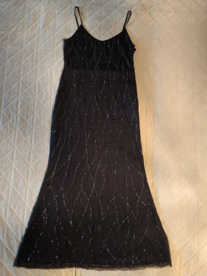 Svart långklänning med pärlor - Svart långklänning med tunna axelband och glittriga pärldetaljer i vågiga mönster över hela klänningen. Klänningen är ca 135 cm lång och har en lätt utsvängd form nedtill. Materialet är tunt och ger ett elegant intryck, perfekt för att sticka ut.