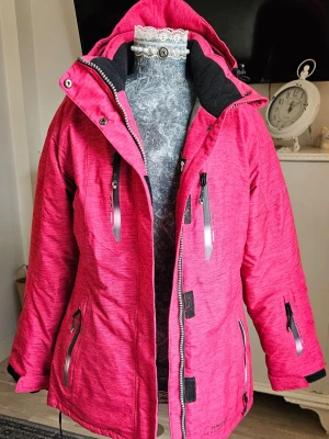 Rosa jacka från Snow Headquarter - Snygg rosa pufferjacka med svart foder och flera praktiska fickor med dragkedja. Jackan har huva, justerbara muddar och detaljer i silver. Perfekt för kalla dagar med sin tjocka och värmande polyester. Stängs med dragkedja och tryckknappar framtill.