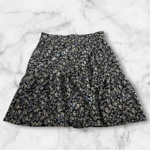 Blommig kortkjol från Lindex - Supersöt kortkjol från Lindex med blommigt mönster i svart, blått och vitt. Kjolen har en volangig nederdel och resår i midjan för extra bekvämlighet. Perfekt för dig som gillar en feminin och lekfull stil.