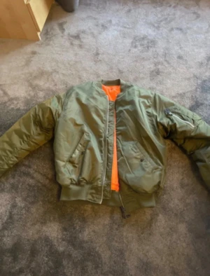 Olivgrön bomberjacka från Brandit - Säljer en olivgrön bomberjacka från Brandit i storlek L. Jackan har orange foder, klassisk ribbad mudd vid ärmslut och midja, samt dragkedja framtill. Materialet är nylon och polyester, med en schysst loose passform och långärmade armar. Perfekt för dig som gillar streetstyle. Originalpris 860