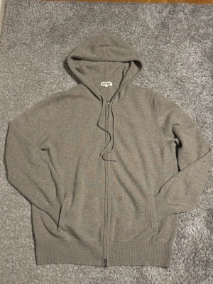 Grå hoodie i kashmir från Soft Goat - Mjuk och stilren grå hoodie från Soft Goat i lyxig kashmir. Tröjan har huva med snörning, hel dragkedja framtill och ribbade muddar vid ärmslut och nederkant. Perfekt för dig som gillar enkel och clean look.