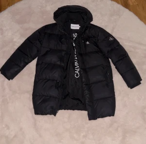 Svart pufferjacka från Calvin Klein - Svart pufferjacka från Calvin Klein Jeans med huva och vita logodetaljer på bröstet och ärmen. Jackan har dragkedja framtill och en extra ficka med zip på bröstet. Materialet är glansigt och syntetiskt, perfekt för kalla dagar.