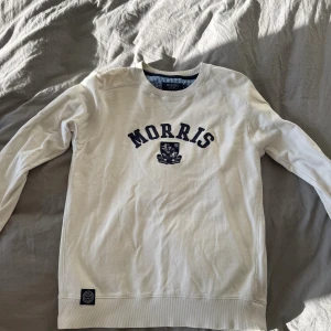 Vit sweatshirt från Morris med tryck - INKÖPT PÅ CAREOFCARL.!                                     Vit sweatshirt från Morris med mörkblå broderad logga och emblem på bröstet. Tröjan har rund halsringning, långa ärmar och ribbade muddar. Tillverkad i mjuk bomull för en skön och avslappnad stil.