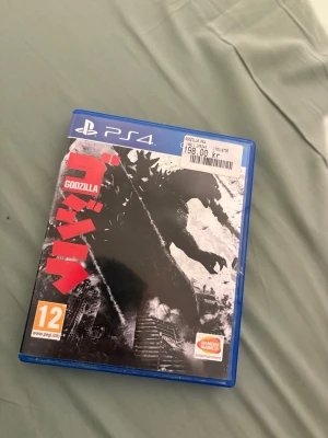 Godzilla PS4 - Godzilla till PlayStation 4, actionfyllt spel där du slåss mot legendariska monster. Fodralet och skivan ser ut att vara i gott skick, med PEGI 12 och stöd för online-spel. Perfekt för fans av monster och film-liknande gameplay.