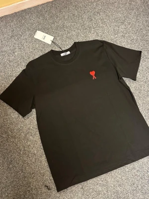 Svart t-shirt från Ami Paris med hjärta - Svart t-shirt från Ami Paris i mjuk bomull. Klassisk rund hals och korta ärmar. På bröstet finns ett broderat rött hjärta med bokstaven A, som ger en snygg detalj. Perfekt för dig som gillar stilrena plagg med en twist.