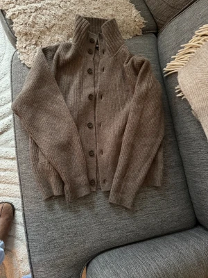 Beige stickad kofta från Uniqlo - Mysig beige stickad kofta från Uniqlo med ribbad struktur och höga muddar. Koftan har hög krage, knappar hela vägen och långa ärmar. Perfekt för lager-på-lager och chill höst- eller vinterstil.