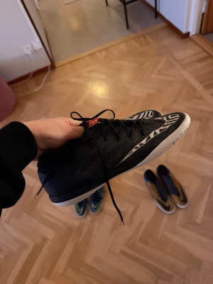Svarta Nike fotbollsskor med mönster - Säljer ett par svarta Nike fotbollsskor med vit sula och coolt grafiskt mönster på sidorna. Skorna har snörning och en röd Nike-logga på plösen och sulan. Perfekta för inomhusspel och har en platt, mönstrad yttersula för bra grepp.