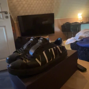 Valentino Garavani sneakers svart/vit - Snygga svarta sneakers från Valentino Garavani med stor vit VLTN-logga på sidan och vit häl. Klassisk rund tå, svarta skosnören och platt sula. Tillverkade i skinn för en lyxig känsla och stilren look. OBS! Skorna har en stor deffekt längst fram på högra skon!!