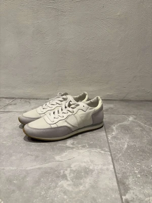 Vita sneakers från philippe model - Snygga vita sneakers med grå mockadetaljer och platt sula. Skorna har rund tå och klassisk snörning. Ovandelen är i en mix av skinn och mocka, vilket ger en stilren och fräsch look. Perfekta för dig som gillar minimalistisk design och neutrala färger. Skorna är i storlek 40