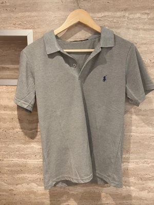 Grå pikétröja från Polo Ralph Lauren - Säljer en stilren grå pikétröja från Polo Ralph Lauren med klassisk krage och korta ärmar. Tröjan har två knappar framtill och den ikoniska blå broderade loggan på bröstet. Perfekt för en clean och chill look tags ingår. Skriv vid frågor