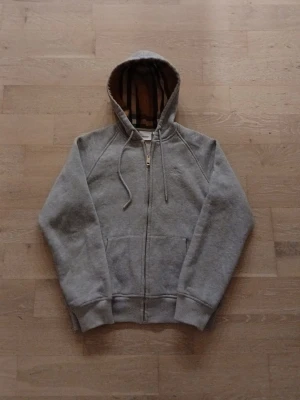 Burberry Zip Hoodie-Grå - Äldre klassisk modell med check-mönstrad luva och broderad logga. Begagnad men i bra skick, inget trasigt och inga fläckar. Kvalité i dragkedja och tyg. Äkta vara.  Kan skickas snabbt. Pris kan diskuteras.      OBS! Storleken är L men passformen är S/M