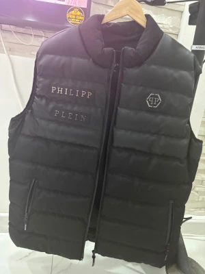 Svart dunväst från Philipp Plein - Svart dunväst från Philipp Plein med quiltad design och logotypdetaljer på bröstet. Den är helt ny och storleken är xl men passar till L oxå. Den är helt ny och it använt den pga att den inte passar mig. 