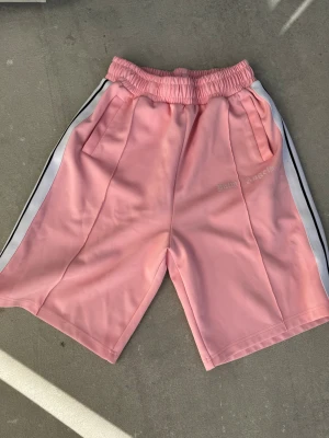 Palm Angels rosa shorts med vita ränder - Säljer ett par snygga rosa shorts från Palm Angels med vita och svarta ränder längs sidorna. De har elastisk midja, fickor och broderad Palm Angels-logga på vänster ben. Perfekt för dig som gillar streetwear och vill sticka ut med färg.