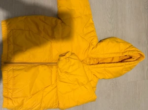 Gul pufferjacka från Zara med huva - Snygg gul pufferjacka från Zara med huva och dragkedja framtill. Jackan har två fickor, elastiska muddar och reflexdetaljer på ena ärmen. Perfekt för kyliga dagar och enkel att matcha med andra plagg. Bra med stark färg på barn! Storleken är 86